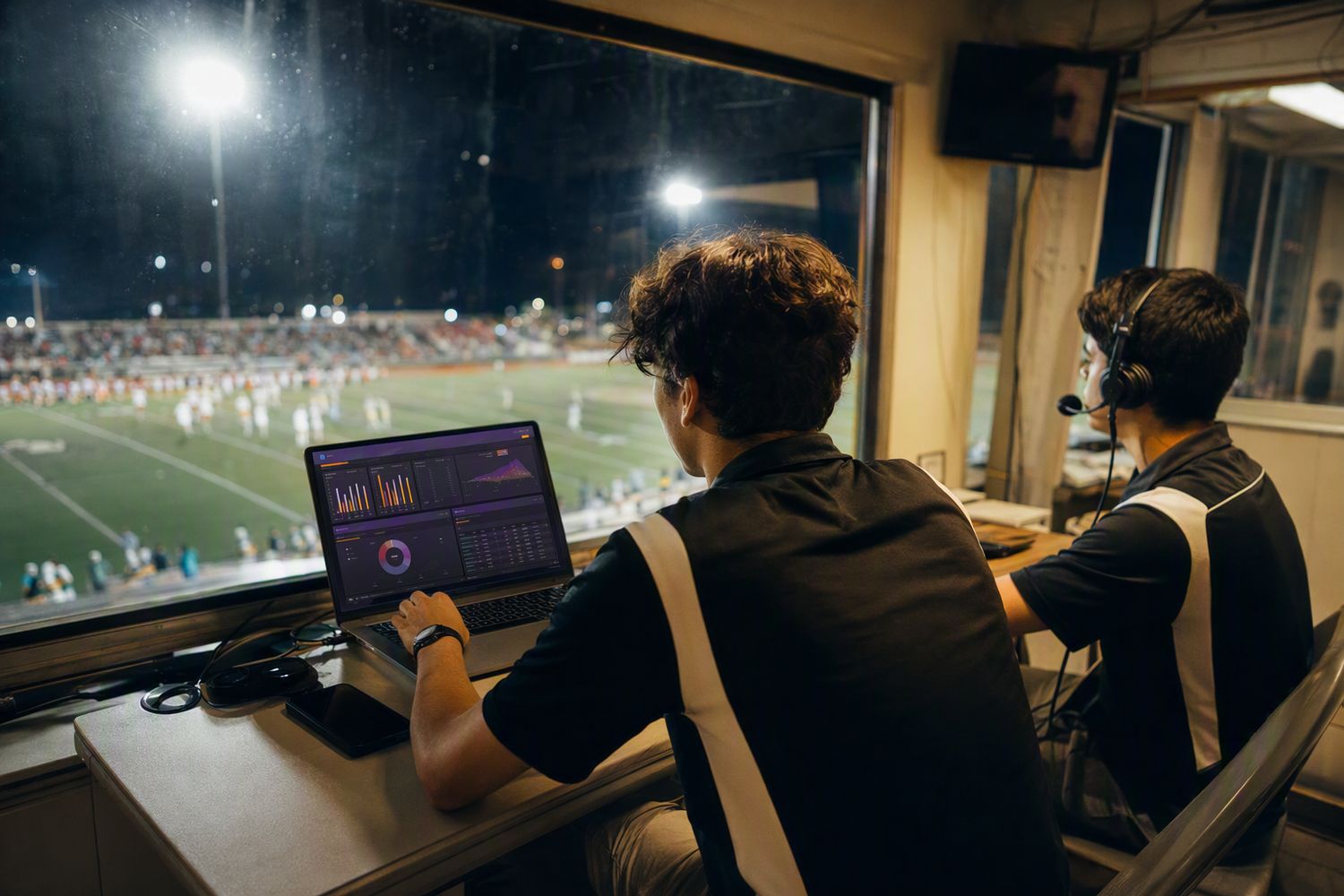 Two student statisticians working in a
high school press box overlooking a floodlit football field